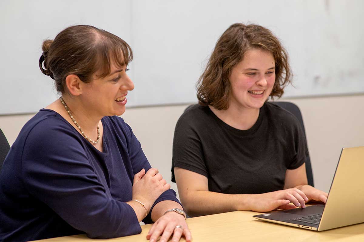 A Khoury professor (left) talks with a student as the student smiles and types on a laptop