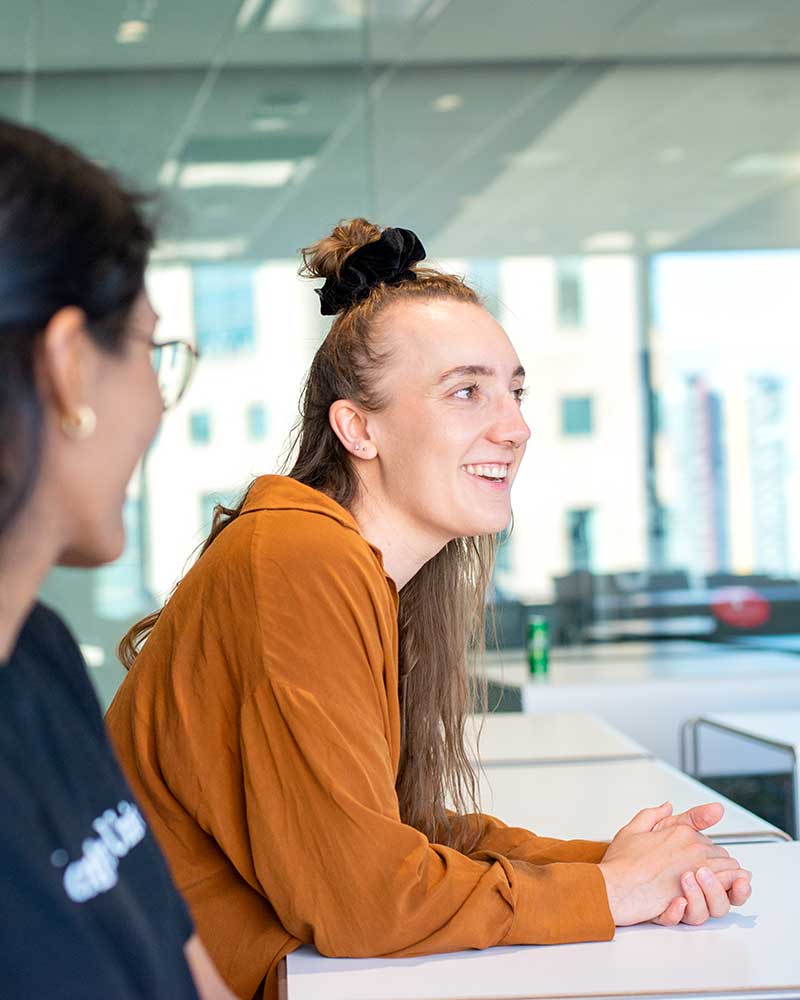 A Khoury student (right) smiles while talking during a class. Another student sits to at a large table to the student's right.