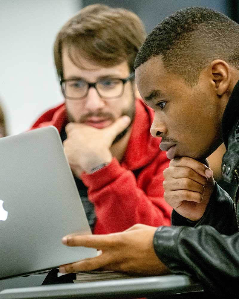 A close photo of two Khoury students looking at an open laptop screen on a desk. Both students are resting their chins on their hands.