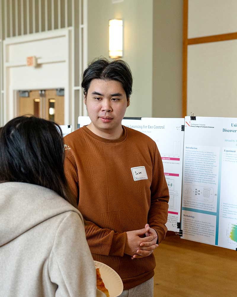 A Khoury student explains his research poster to another student during a research fair