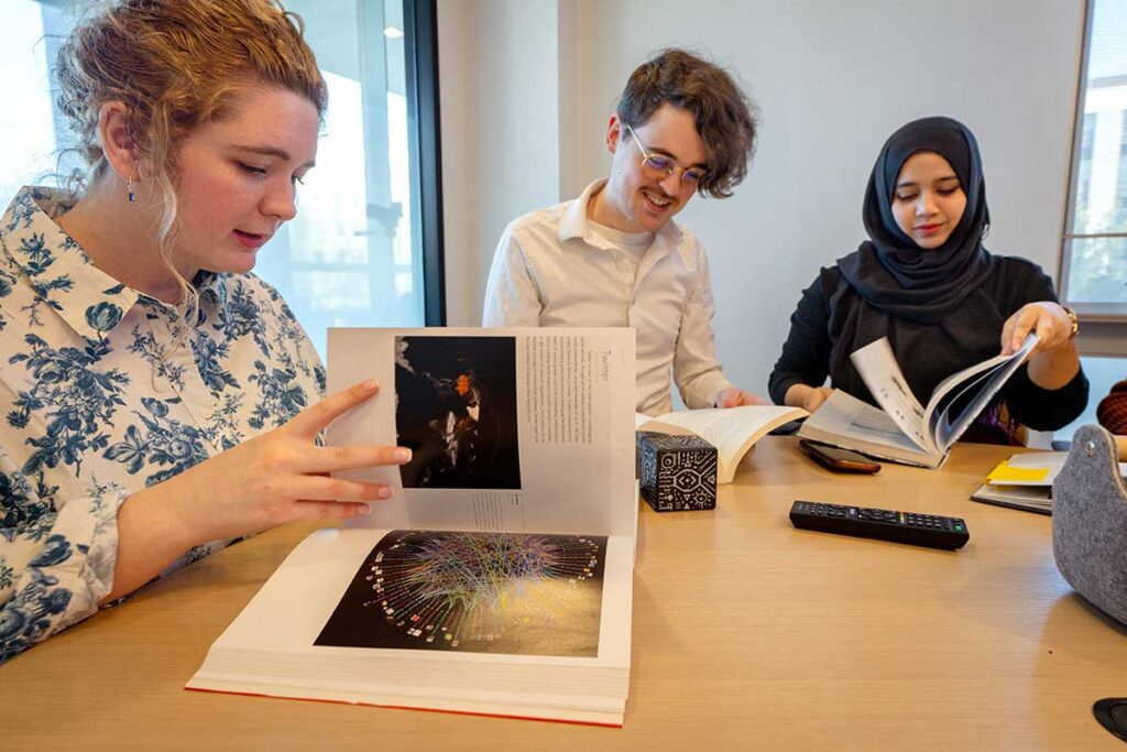 Three Khoury PhD students sit at a large table flipping through data visualization books