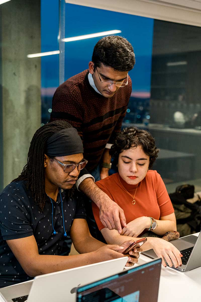 Two students sit at a desk looking at a mobile device while a faculty member stands behind them and points at the screen.