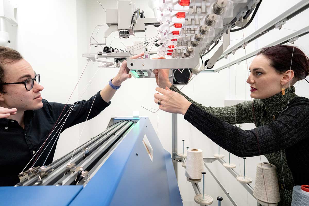 Khoury faculty Megan Hofmann (right) adjusts thread in a knitting machine that knits using artificial intelligence. Another research adjusts the machine.