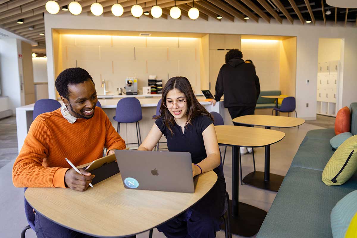 Two students sit around a table viewing a laptop. One students is holding a tablet.