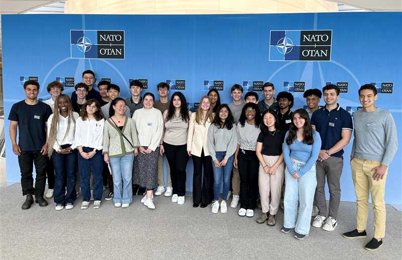 Dozens of students stand in front of a NATO banner