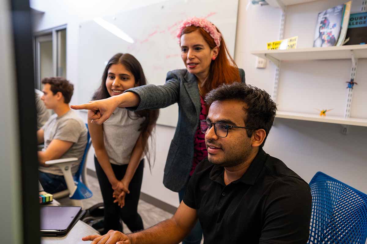 A teacher in the company of students points to a computer monitor.