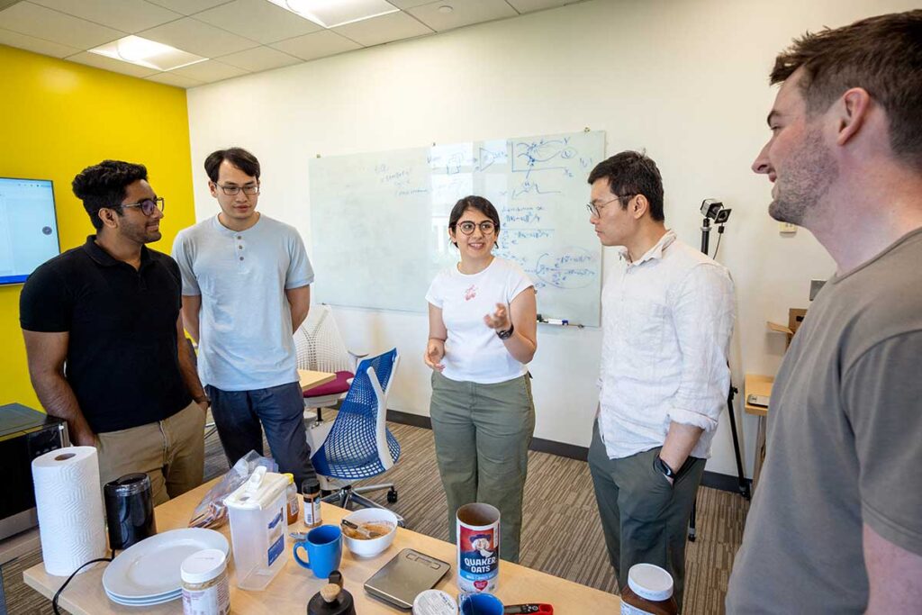 Five Khoury students discuss research in a Khoury lab. A whiteboard covered in writing is in the background.