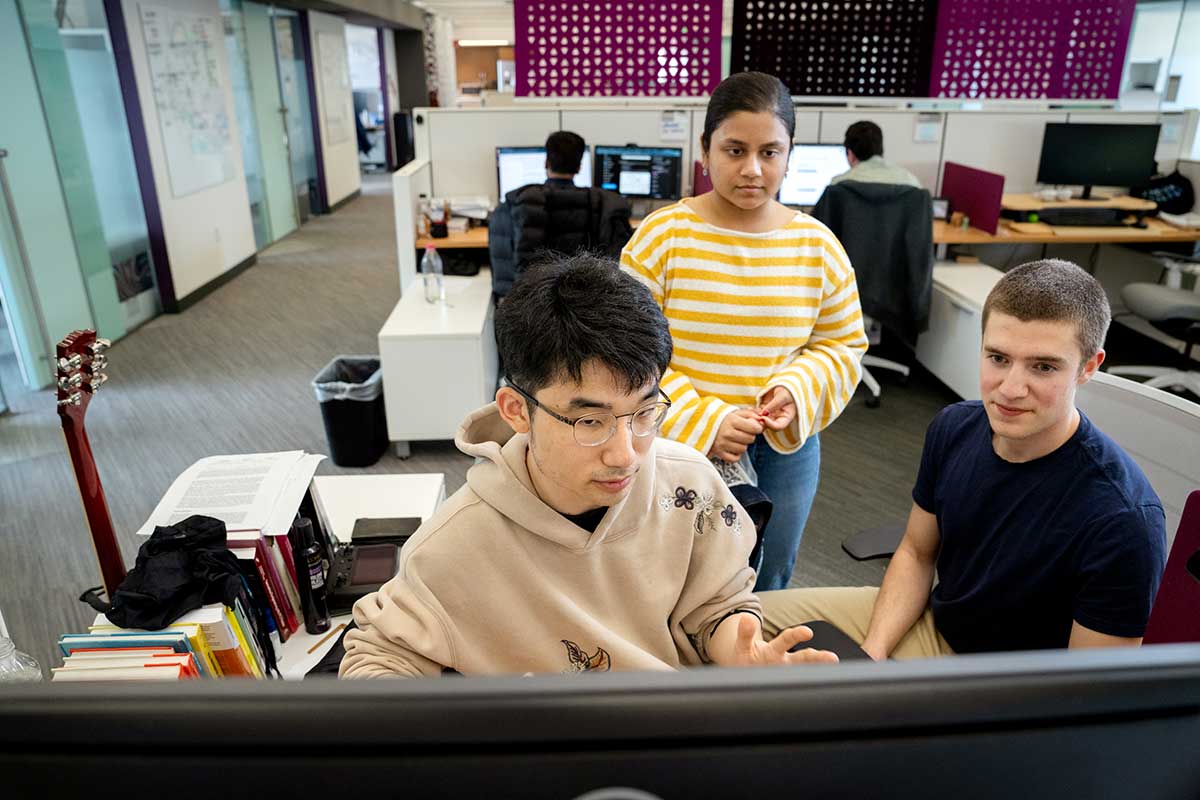 Two students sit at a desk with another student standing behind them. They are all looking at a computer screen.