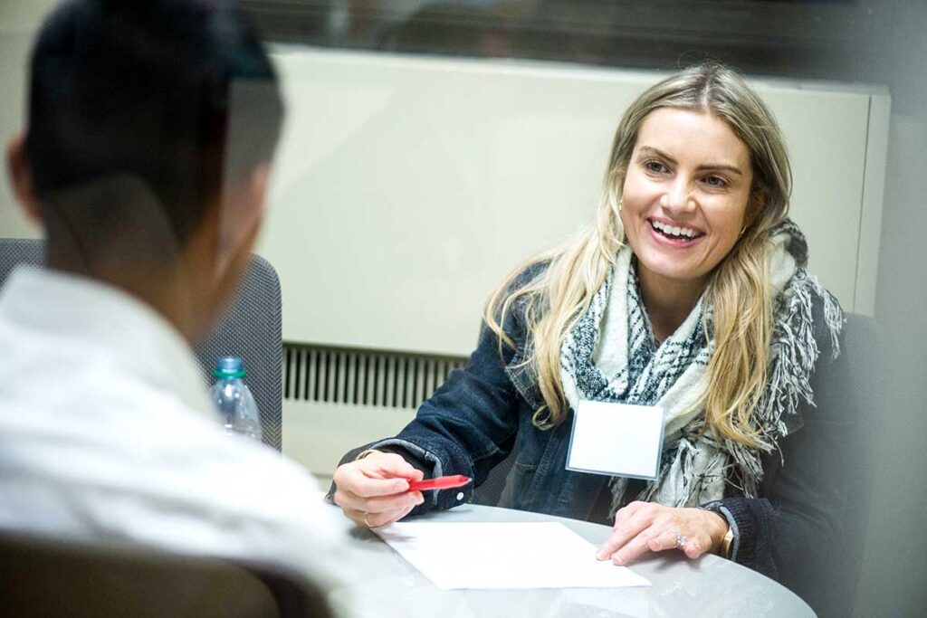 A women sitting on one side of the table holds a piece of paper while talking to a student