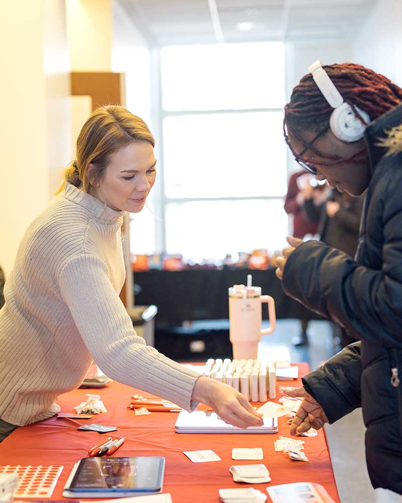 A Khoury staff member stands on one side of a red table and hands a name tag to a student attending an event