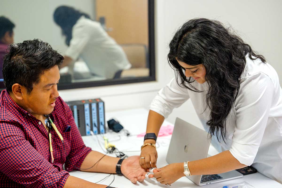 Khoury personal health informatics researchers work in a lab. One researcher connects a wired device to her colleague's finger.