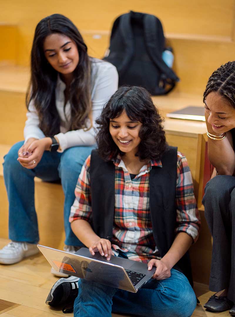 Three students sit on wooden steps; the student in the center is smiling with a laptop open on her lap