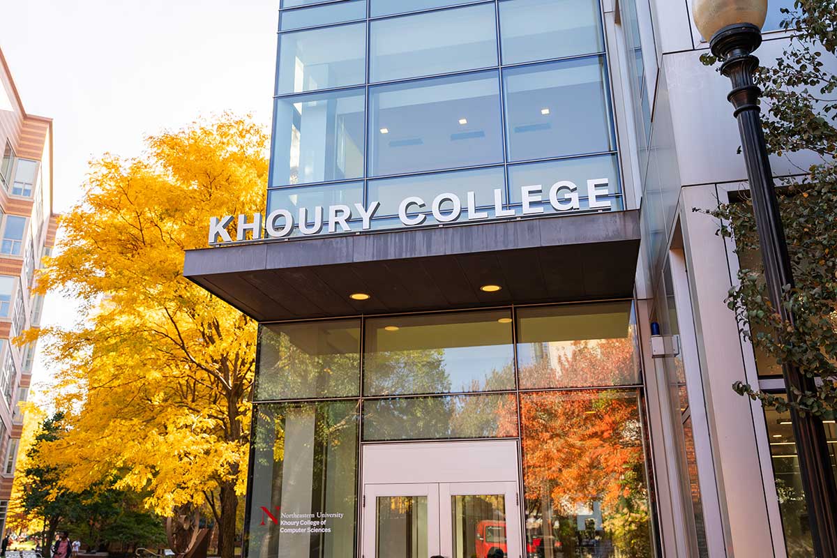 The Khoury College sign at the entrance of West Village H on Northeastern's Boston campus. Yellow fall foliage is visible in the background and reflecting off the building's exterior.