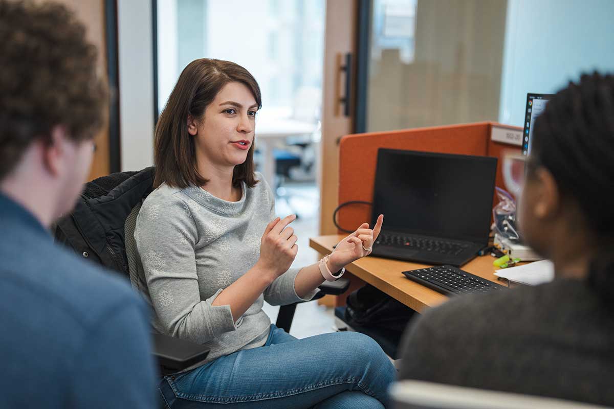 A Khoury student sitting a desk in an office describes her research to two other students