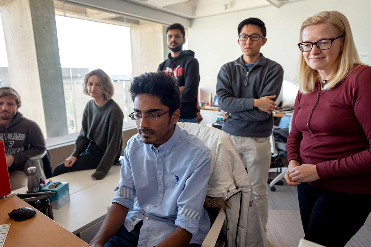 A Khoury student sitting at a desk works on a computer while fellow Khoury researchers stand behind him and look over his shoulder.