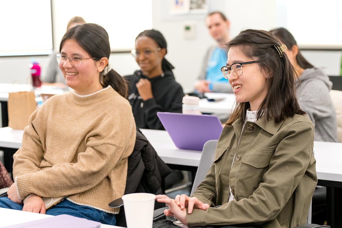 Two students sitting at a long table smile during a course lecture. Other students sit at rows of tables in the background.