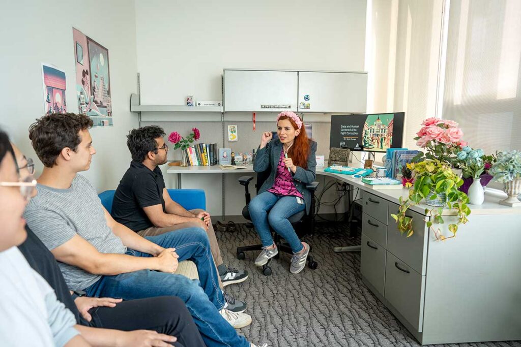 Khoury faculty member Saiph Savage sits in a chair in her office and speaks to four students sitting on sofas