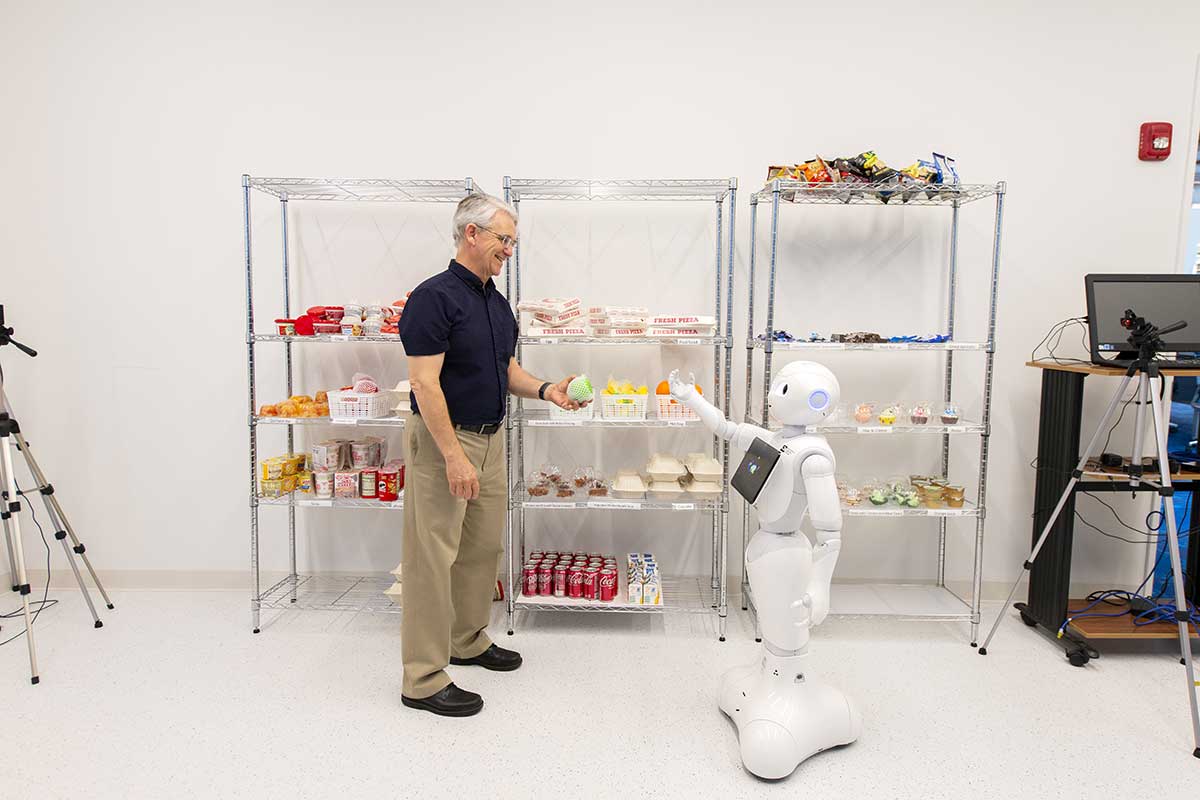 Khoury faculty member Timothy Bickmore hands an object to a robot shaped like a human. The robot's arm is outstretched.