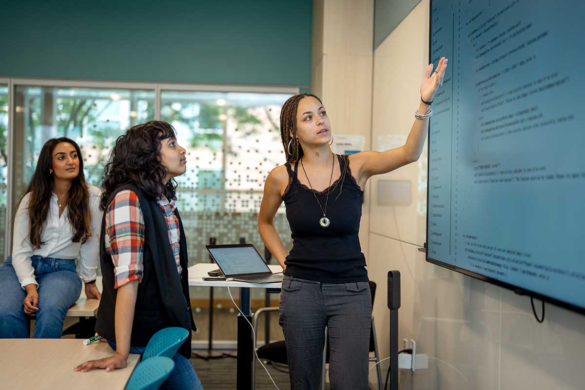 A Khoury student points at text projected on a large screen in a conference room while two other students look on