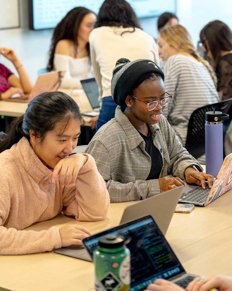 Two students sit at a large table and work on their laptops while talking