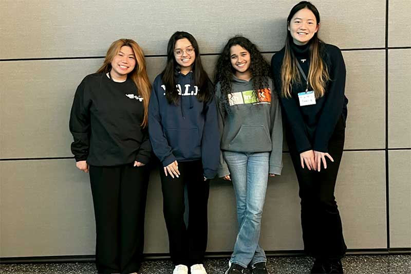 Shannen Espinosa (left) smiles standing next to three other students in a hallway at the iQuHACK event