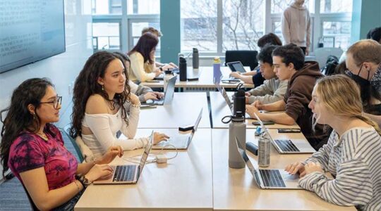 Several students work on a project during a class while sitting around four large tables pushed together