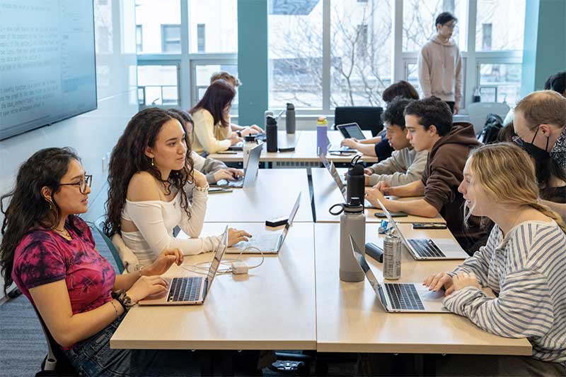 Several students work on a project during a class while sitting around four large tables pushed together