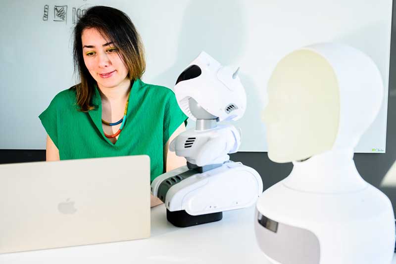 Malihe Alikhani sits at a desk typing on a laptop. Two robotic devices are standing on the desk.