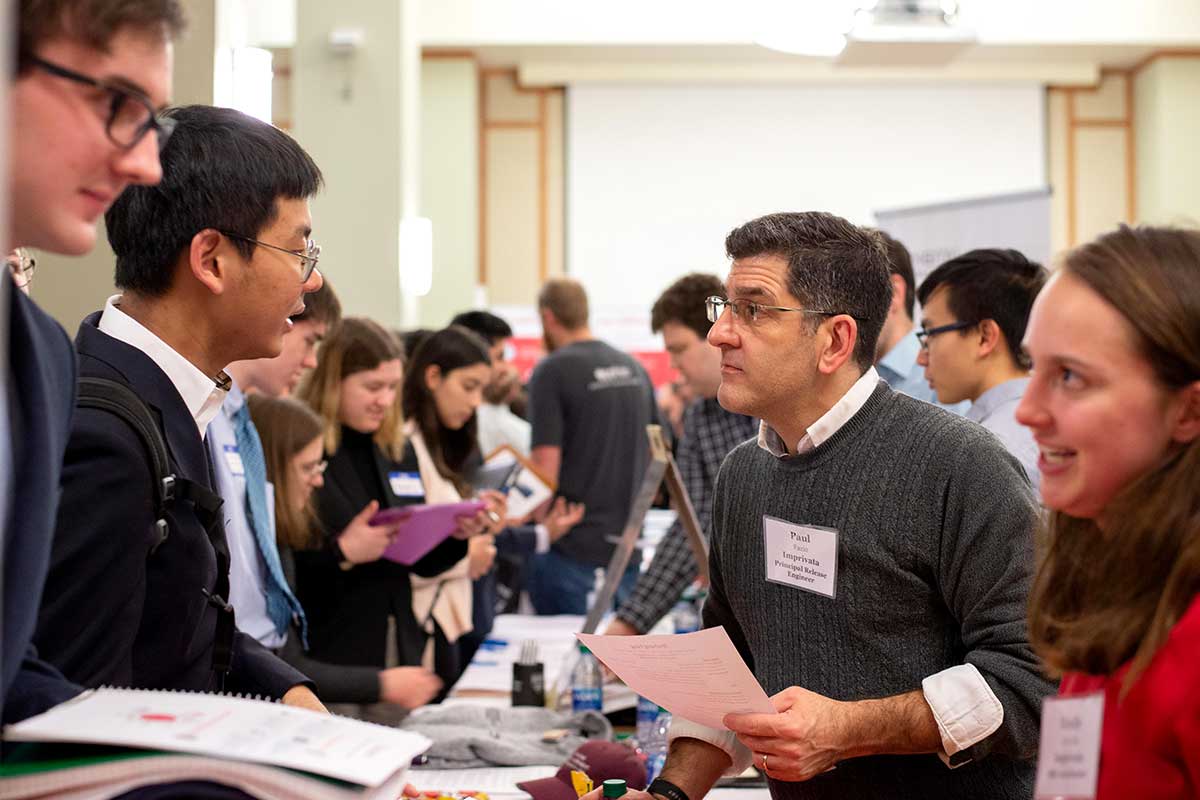 Two employers stand on the right side of a long table while answering questions from students standing on the other side of the table