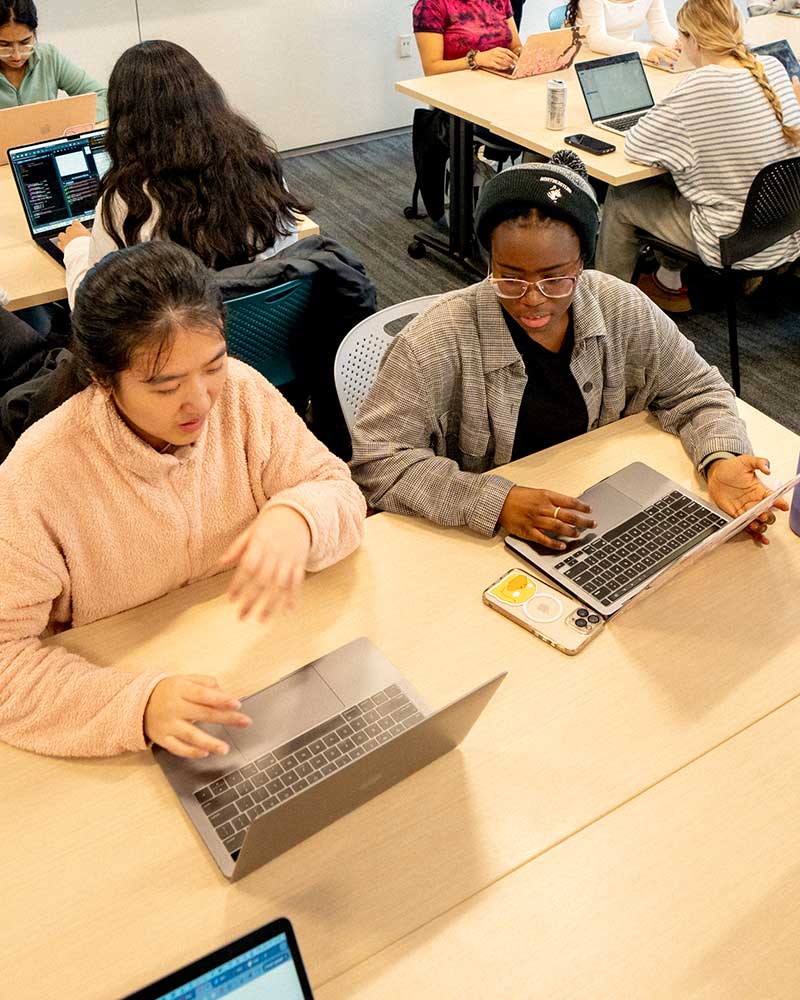 Two students sitting at a rectangular table with open laptops discuss a project while sitting in a Khoury classroom