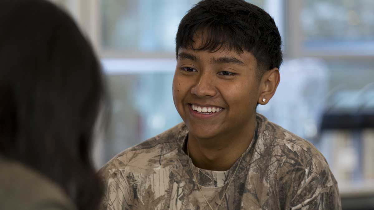 In Seattle, one student sitting at the far side of a table smiles while talking to someone sitting on the other side of the table.