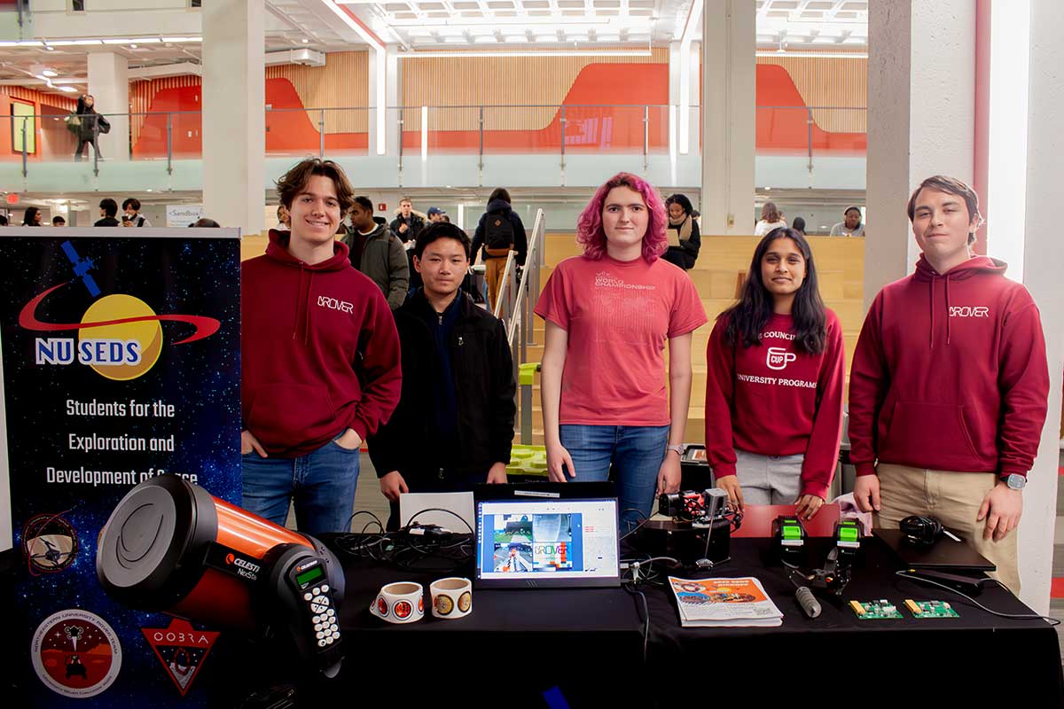 Five members of Students for the Exploration and Development of Space stand behind the club's table at the Northeastern club fair. There is a telescope, open laptop, and rolls of stickers on the club's table.