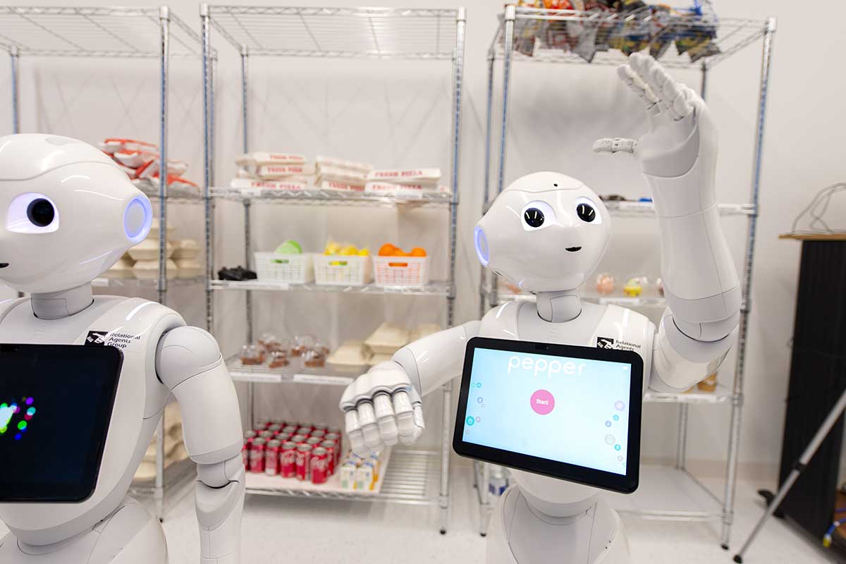 A photo of two robots in a Khoury lab. The robots resemble human bodies, including heads with eyes and a mouth. The robots have tablet devices on their chests.