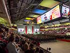 A photo from the back of Matthews Arena shows a large crowd watching Dean Mynatt's address, which is also visible on the video screen hanging from the ceiling