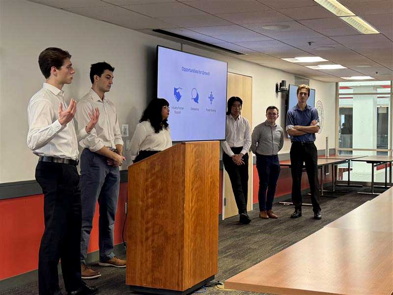 The NExT team presents a slideshow standing behind a podium adjacent to a large screen