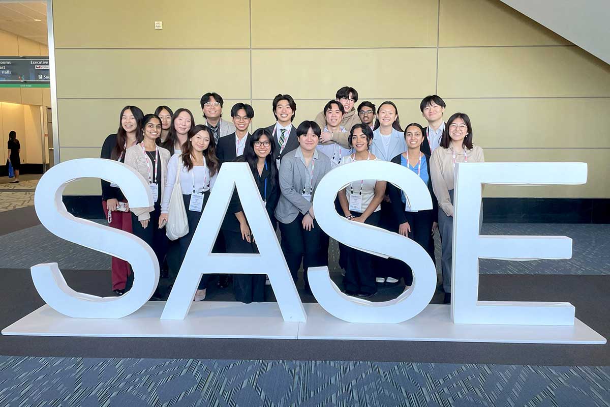 18 members of the Society of Asian Scientists and Engineers poses behind a SASE sign in a conference hall