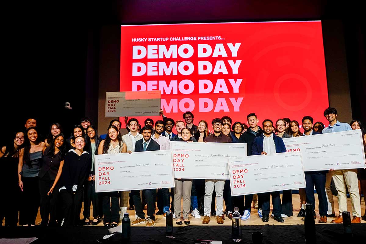Dozens of award winners stand on a stage holding large novelty checks that say "DEMO DAY FALL 2024"