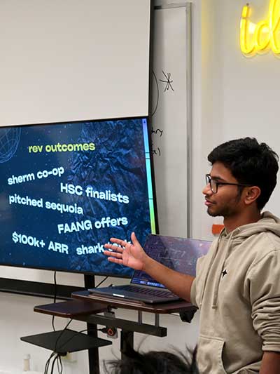 A member of Rev gestures at a presentation screen during a meeting. The screen shows multiple outcomes of Rev membership.