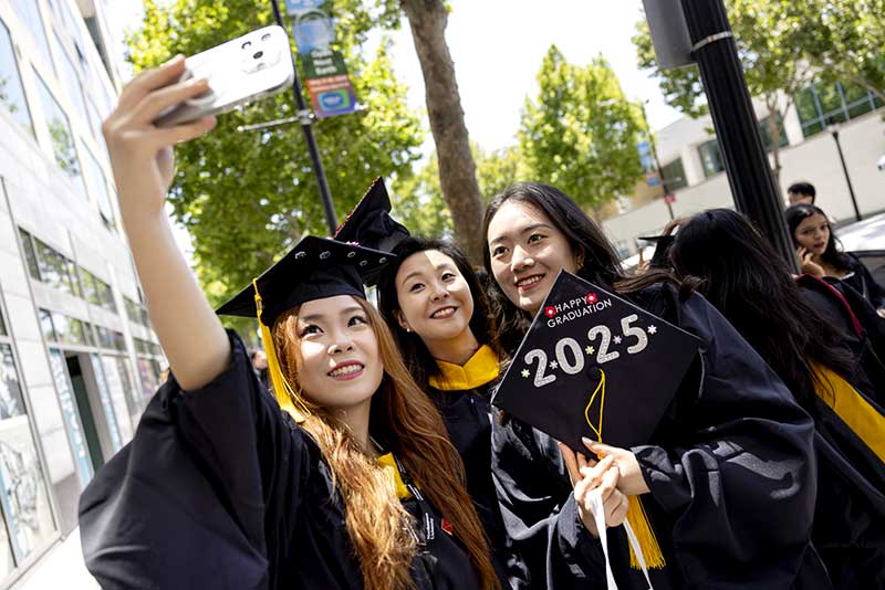 Three graduates pose for a selfie. One graduate holds a cap in her hands that is decorated with the words "Happy graduation 2025"