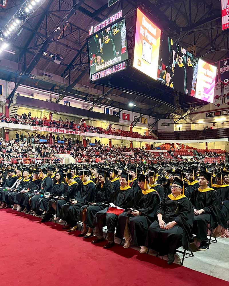 Dozens of rows of graduates in dressed in caps and gowns watch the stage