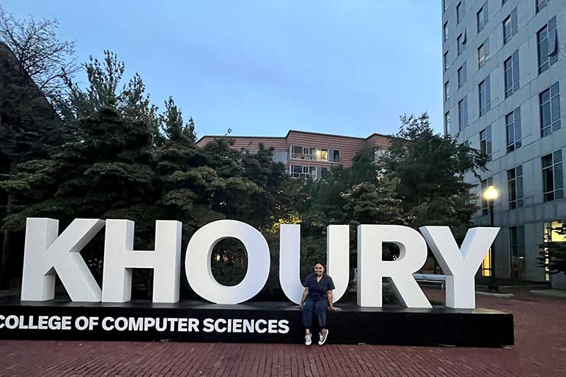 Marcela Perro sits on a large Khoury College of Computer Sciences sign outside West Village H on the Boston campus