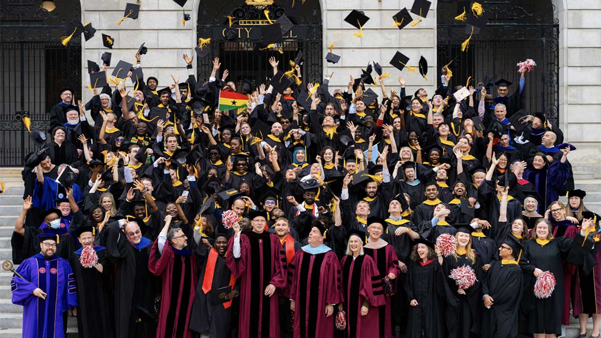 The graduating class of the Roux institute stands on the steps of a building and toss their graduation caps in the air