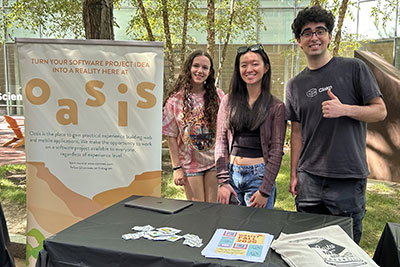 Three students from the oasis student club pose for a photo standing next to their club's poster
