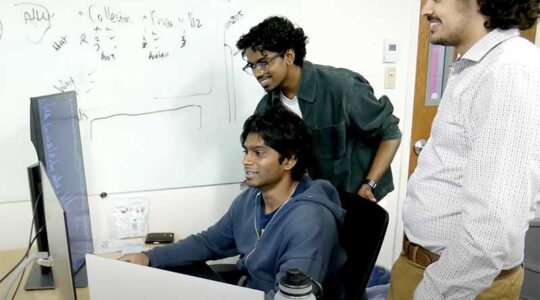 A co-op consultancy student sits at a desk viewing a screen while two members of his group stand behind him