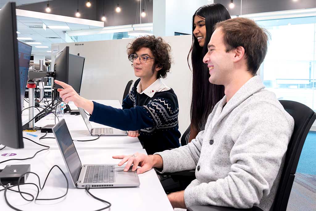 Three Khoury students work on a project in a large office. One student points at a computer screen.