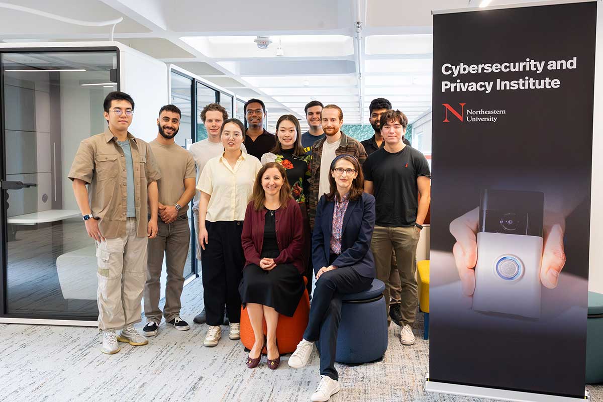 A group of cybersecurity researchers including Khoury faculty Alina Oprea and Cristina Nita-Rotaru pose for a photo in a lab next to a Cybersecurity and Privacy Institute banner