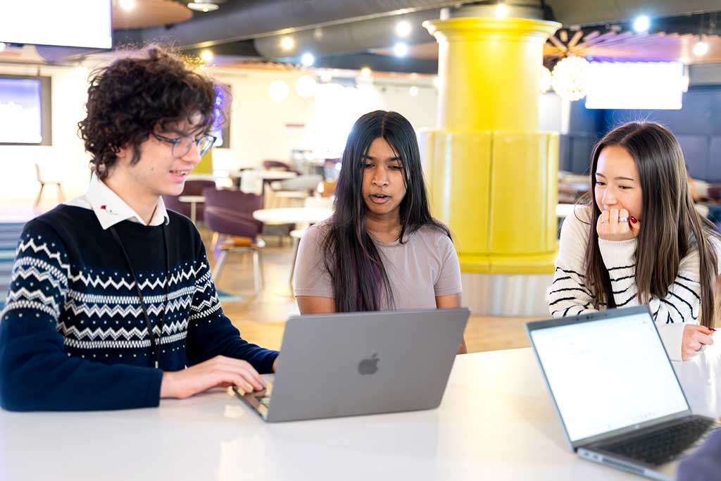 Three Khoury students sit around a large table and talk about a project as one student types on a laptop.