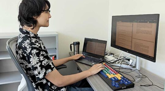 Erika Melder sits at a desk with a laptop open and a larger keyboard and monitor adjacent to the laptop.