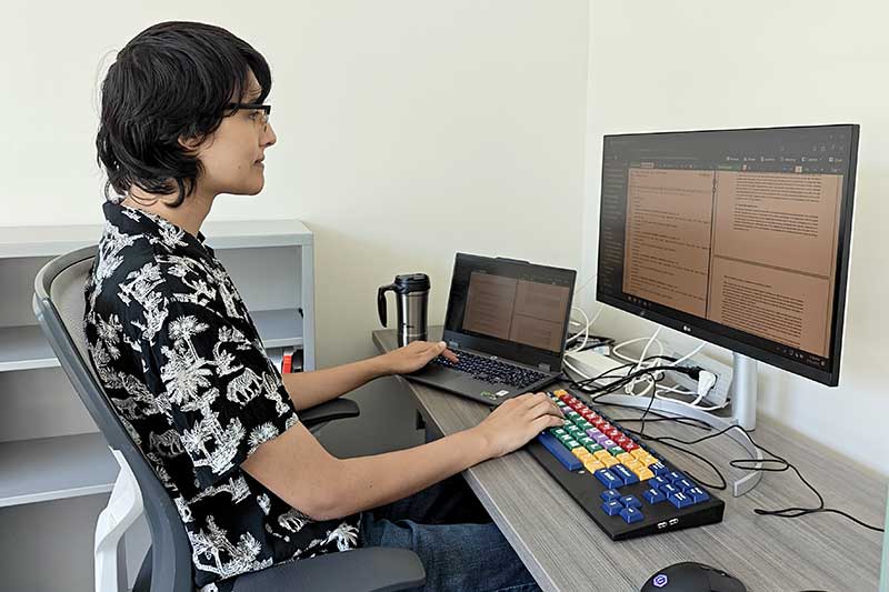 Erika Melder sits at a desk with a laptop open and a larger keyboard and monitor adjacent to the laptop.