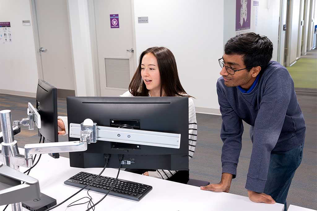 A Khoury student sitting at a desk with a keyboard and two monitors points at the right monitor while a colleague looks on